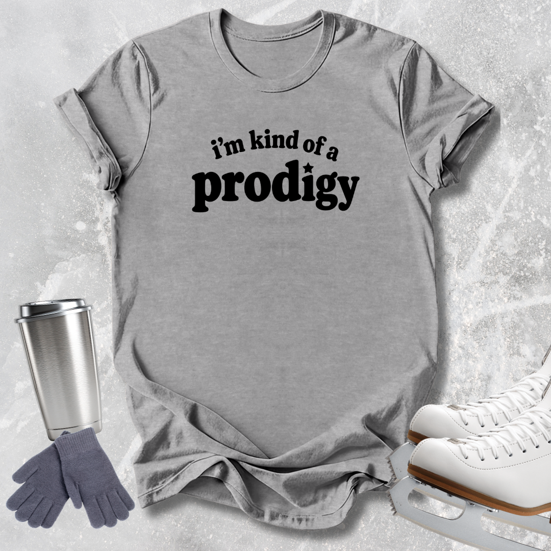 A heather gray short-sleeve t-shirt with the black text 'i'm kind of a prodigy' printed on the chest, styled in a flat lay with ice skates, gloves, and a travel tumbler on a light gray textured background.