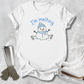 A white short-sleeve T-shirt laid flat on a light gray textured surface, featuring a cartoon snowman wearing a blue beanie and scarf with the text 'I'm melting' above it. A stainless steel travel mug, gray gloves, and white ice skates are arranged around the shirt as props.
