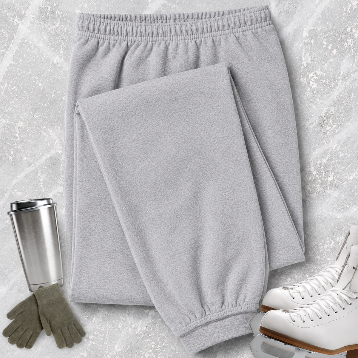 Light gray fleece sweatpants laid flat on a textured light gray surface, styled with a stainless steel travel mug, gloves, and white ice skates.
