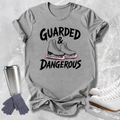 A heather gray graphic t-shirt featuring the text 'GUARDED & DANGEROUS' in black with an illustration of ice skates in the center. The shirt is styled in a flat-lay arrangement with a stainless steel travel mug, gray gloves, and white ice skates on a light gray textured background.