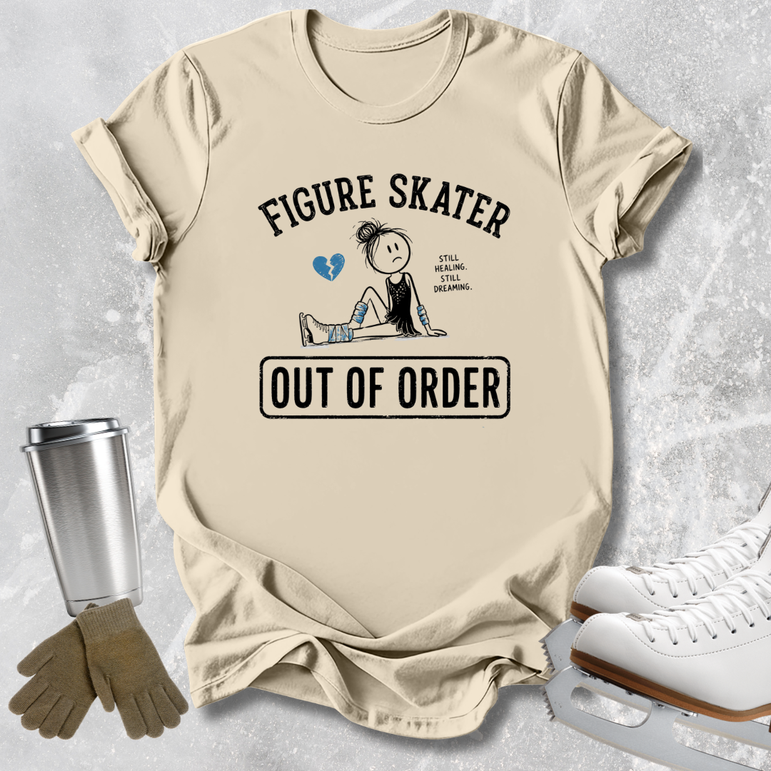 A beige graphic t-shirt laid flat on a light gray textured surface, featuring a figure skater illustration with the text 'FIGURE SKATER OUT OF ORDER' and a small blue heart. White ice skates, tan gloves, and a metal travel tumbler are arranged around the shirt as styling props.