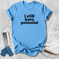 A light blue short-sleeve T-shirt with the phrase 'i still have potential' printed in black across the chest, styled in a flat lay with ice skates, gloves, and a travel tumbler on a light gray textured surface.
