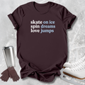 A dark brown short-sleeve graphic T-shirt laid flat on a light gray textured surface, with the phrase 'skate on ice spin dreams love jumps' printed in white and light blue text. A stainless steel travel mug, brown gloves, and white ice skates are arranged around the shirt as styled props.