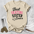 A beige graphic T-shirt laid flat on a light gray textured surface, with the text 'Best SKATING Sister Ever' in black and pink lettering and a small ice skate illustration. Ice skates, gloves, and a metal cup are styled around the shirt as props.