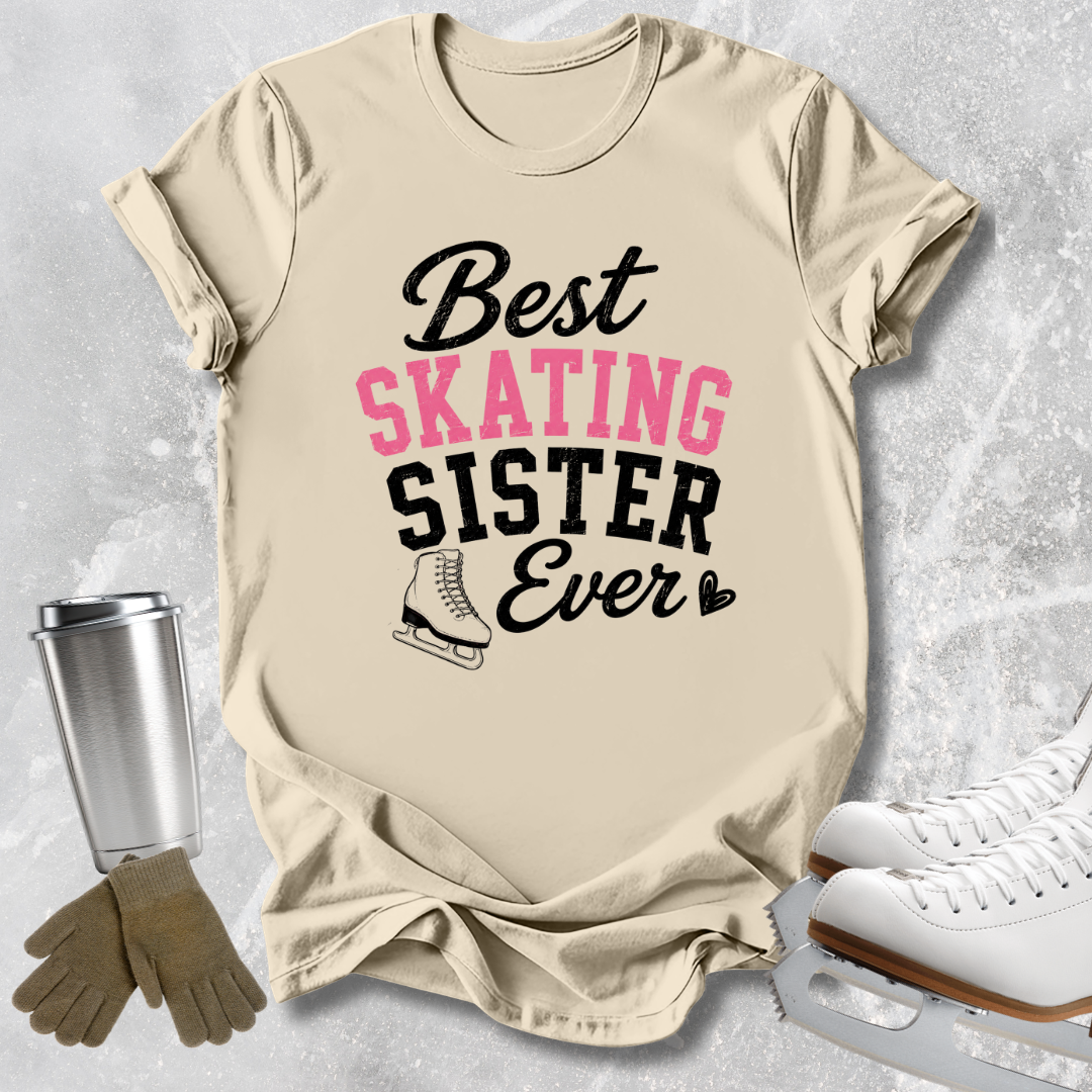 A beige graphic T-shirt laid flat on a light gray textured surface, with the text 'Best SKATING Sister Ever' in black and pink lettering and a small ice skate illustration. Ice skates, gloves, and a metal cup are styled around the shirt as props.