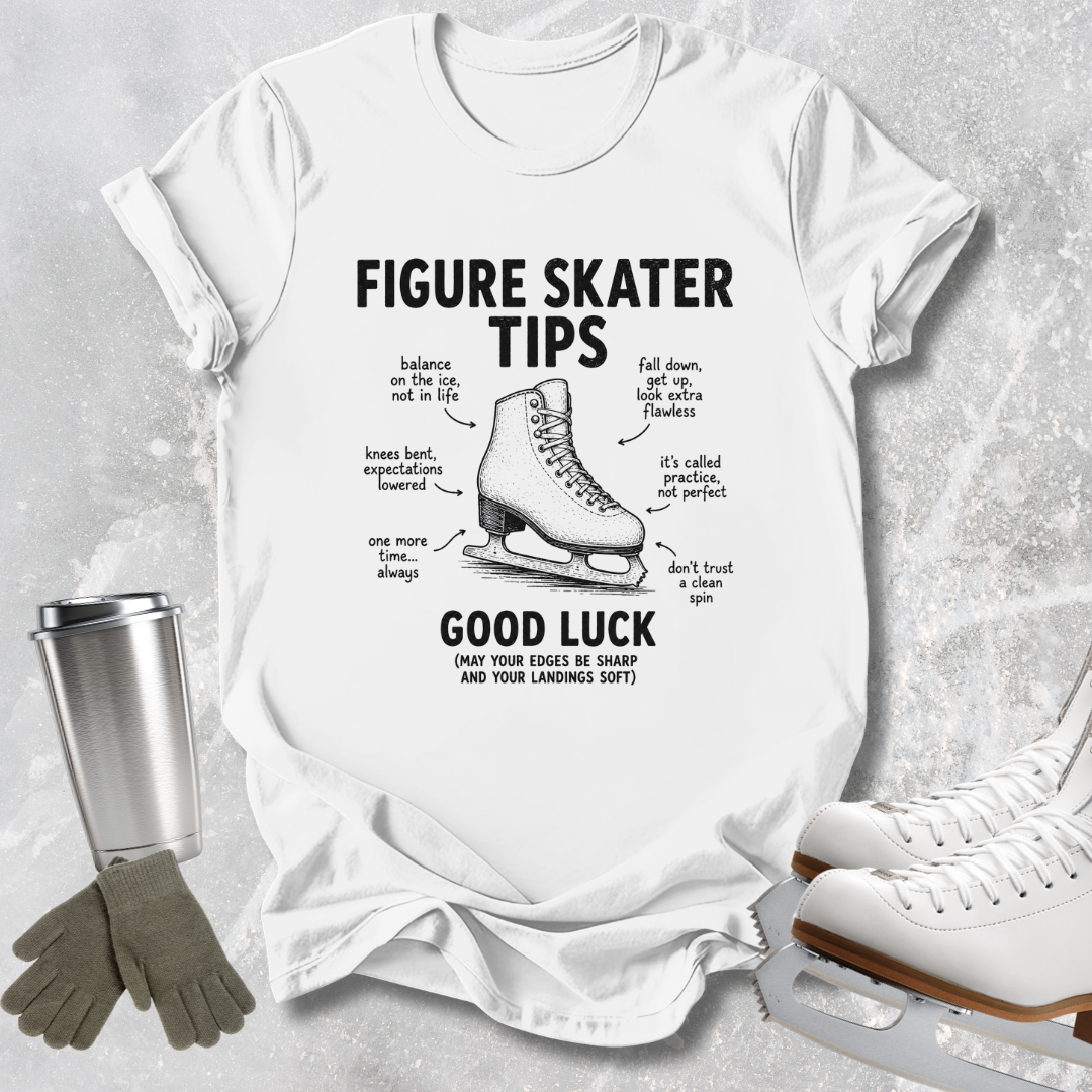 A white graphic t-shirt featuring a black illustration of a figure skate and humorous tips labeled 'FIGURE SKATER TIPS' and 'GOOD LUCK'. The shirt is styled in a flat-lay arrangement on a textured gray background, accompanied by a silver travel mug, gray gloves, and white ice skates.