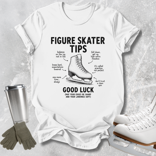 A white graphic t-shirt featuring a black illustration of a figure skate and humorous tips labeled 'FIGURE SKATER TIPS' and 'GOOD LUCK'. The shirt is styled in a flat-lay arrangement on a textured gray background, accompanied by a silver travel mug, gray gloves, and white ice skates.
