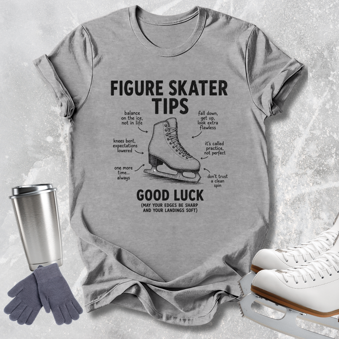 A light gray graphic t-shirt featuring a black print of a figure skate with the text 'FIGURE SKATER TIPS' and 'GOOD LUCK'. The shirt is displayed in a flat-lay arrangement on a textured gray background, accompanied by a silver travel mug, gray gloves, and white ice skates.