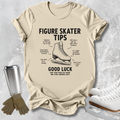 A beige graphic t-shirt featuring a black print of a figure skater boot with handwritten-style tips and notes around it. The shirt is displayed in a flat-lay arrangement on a textured gray background, accompanied by a silver travel mug, brown gloves, and white ice skates.