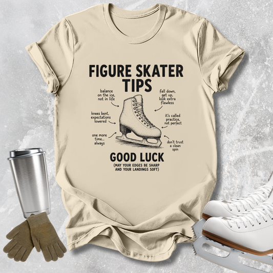 A beige graphic t-shirt featuring a black print of a figure skater boot with handwritten-style tips and notes around it. The shirt is displayed in a flat-lay arrangement on a textured gray background, accompanied by a silver travel mug, brown gloves, and white ice skates.
