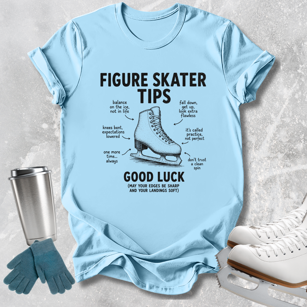 A light blue graphic T-shirt laid flat on a textured light gray surface, with black text reading 'FIGURE SKATER TIPS' and 'GOOD LUCK' around an illustration of a figure skate boot. A stainless steel cup, blue gloves, and white ice skates are placed around the shirt as props.