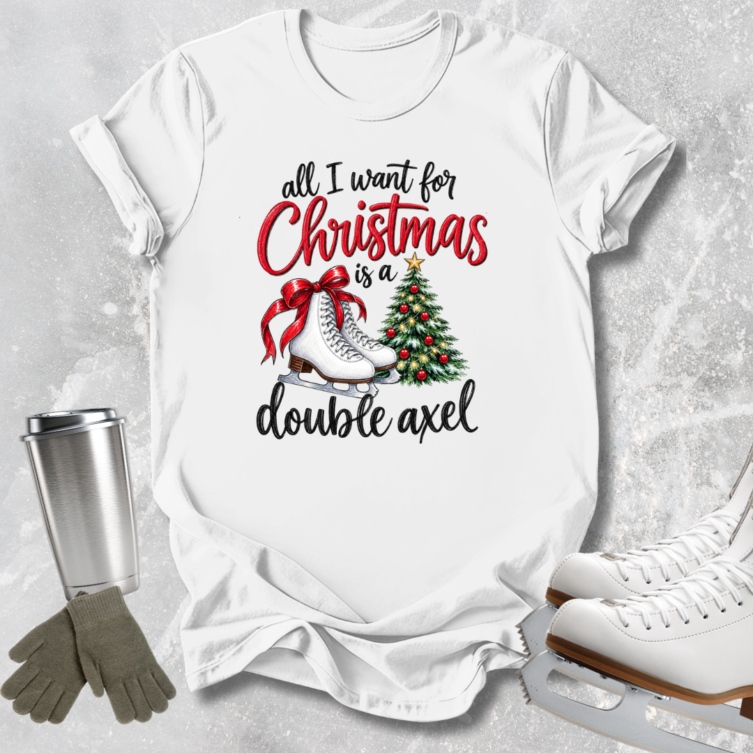 A white graphic t-shirt laid flat on a light gray textured surface, featuring the text 'all I want for Christmas is a double axel' in black and red lettering, with an illustration of ice skates, a red bow, and a Christmas tree. A stainless steel tumbler, gray gloves, and white ice skates are styled around the shirt.