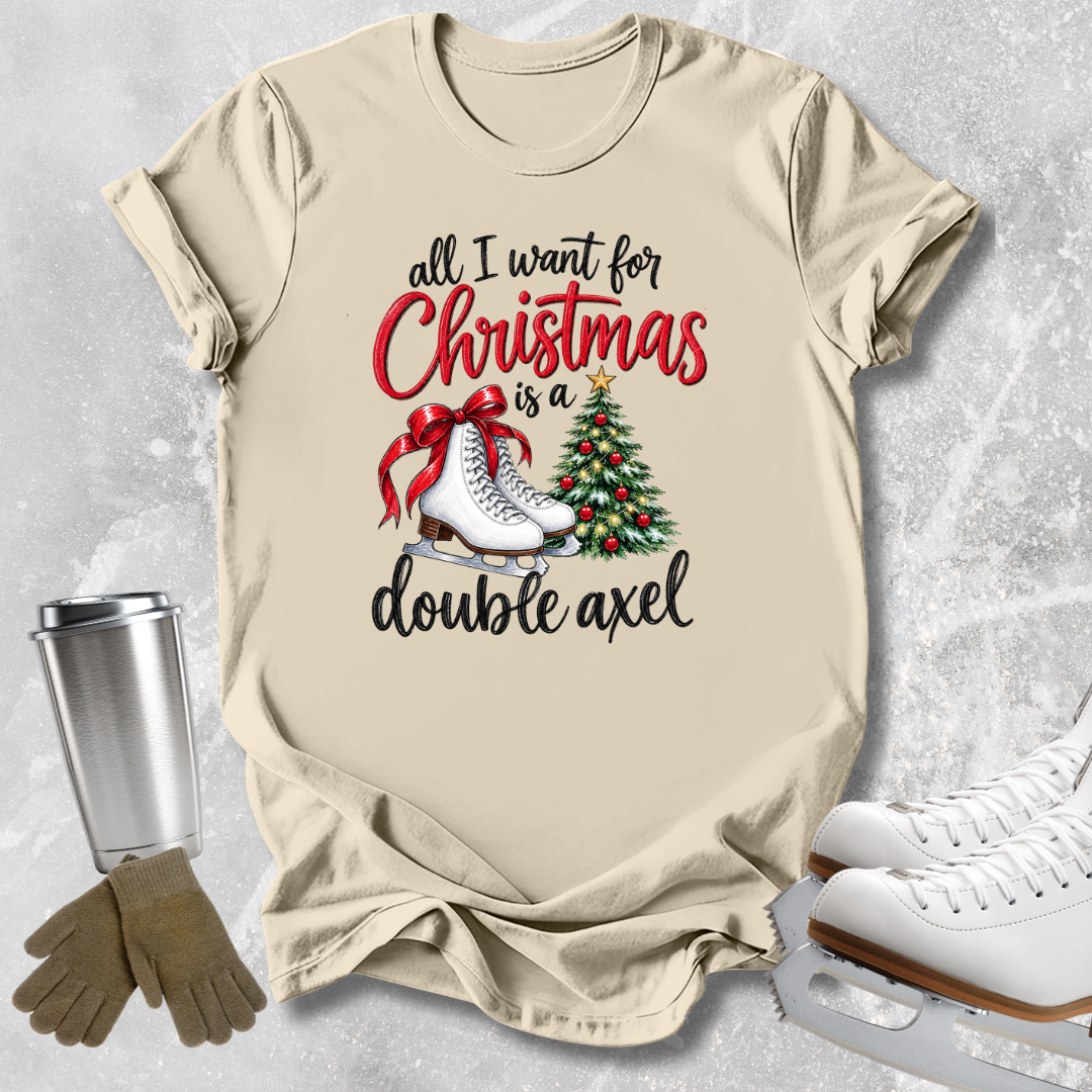 A beige graphic T-shirt laid flat on a light gray textured surface, with a Christmas-themed print reading 'all I want for Christmas is a double axel' in black and red lettering, plus skates, a tree, and a red bow. A metal tumbler, gloves, and white ice skates are arranged around the shirt as styling props.