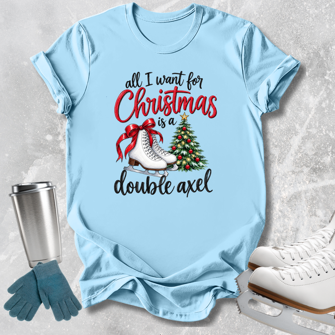 A light blue graphic T-shirt laid flat on a textured light gray surface, with a Christmas-themed print reading 'all I want for Christmas is a double axel' in black and red lettering, plus skates, a red bow, and a decorated tree. A metal tumbler, blue gloves, and white ice skates are arranged around the shirt as props.