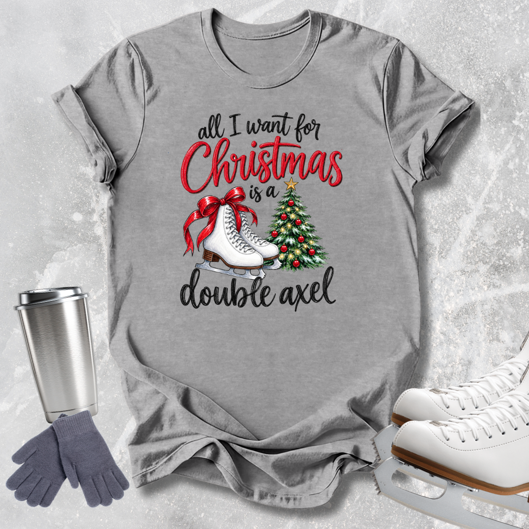 A heather gray graphic T-shirt laid flat on a light textured surface, featuring the phrase 'all I want for Christmas is a double axel' in black and red lettering, with white ice skates and a decorated Christmas tree illustration. A stainless steel tumbler, gray gloves, and white ice skates are arranged around the shirt as styled props.