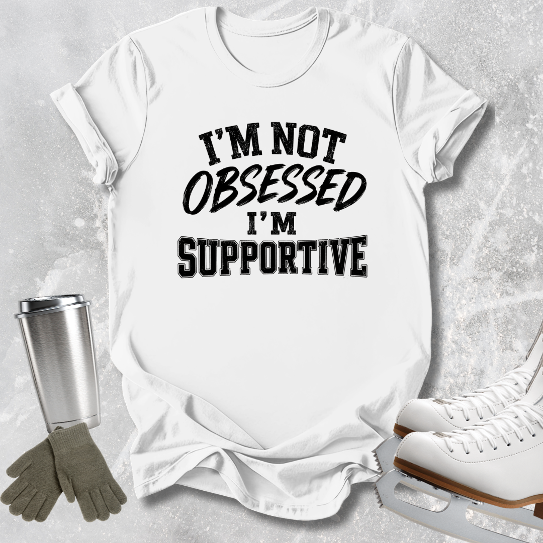 A white short-sleeve graphic T-shirt laid flat on a light gray textured surface, with black text reading “I'M NOT OBSESSED I'M SUPPORTIVE.” Ice skate boots, gloves, and a metal cup are arranged around it as styling props.