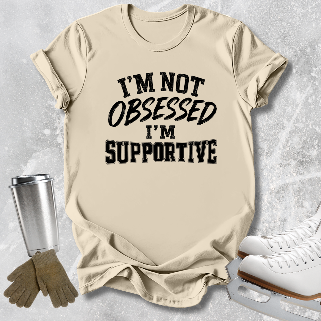 A beige graphic t-shirt with the text 'I'M NOT OBSESSED I'M SUPPORTIVE' in black, styled in a flat-lay with ice skates, gloves, and a travel tumbler on a gray textured background.