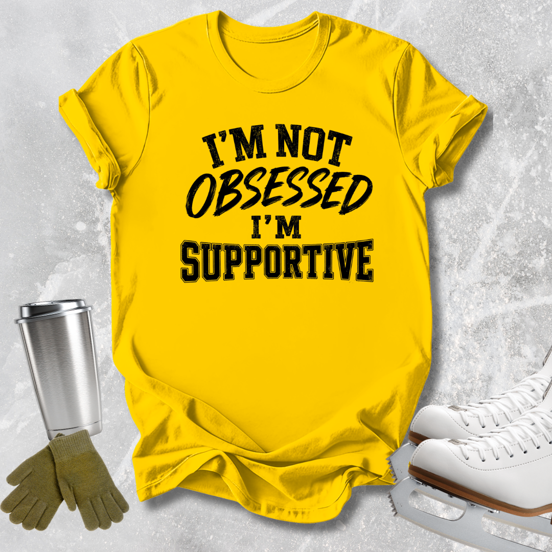 A bright yellow graphic t-shirt with the text 'I'M NOT OBSESSED I'M SUPPORTIVE' in black, styled in a flat-lay with ice skates, gloves, and a metal cup on a light gray textured background.