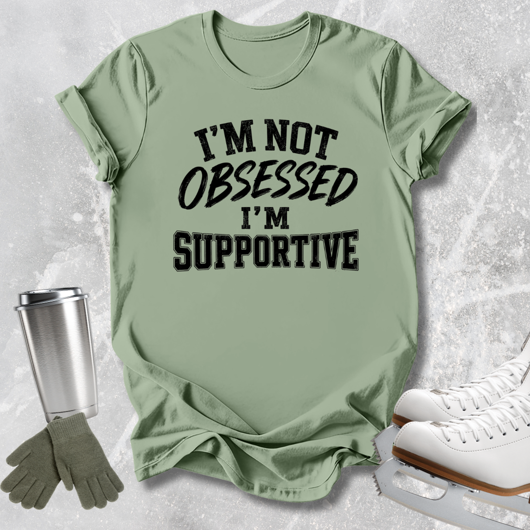 A sage green graphic t-shirt with the text 'I'M NOT OBSESSED I'M SUPPORTIVE' in black, styled in a flat-lay with ice skates, gloves, and a travel tumbler on a light gray textured background.