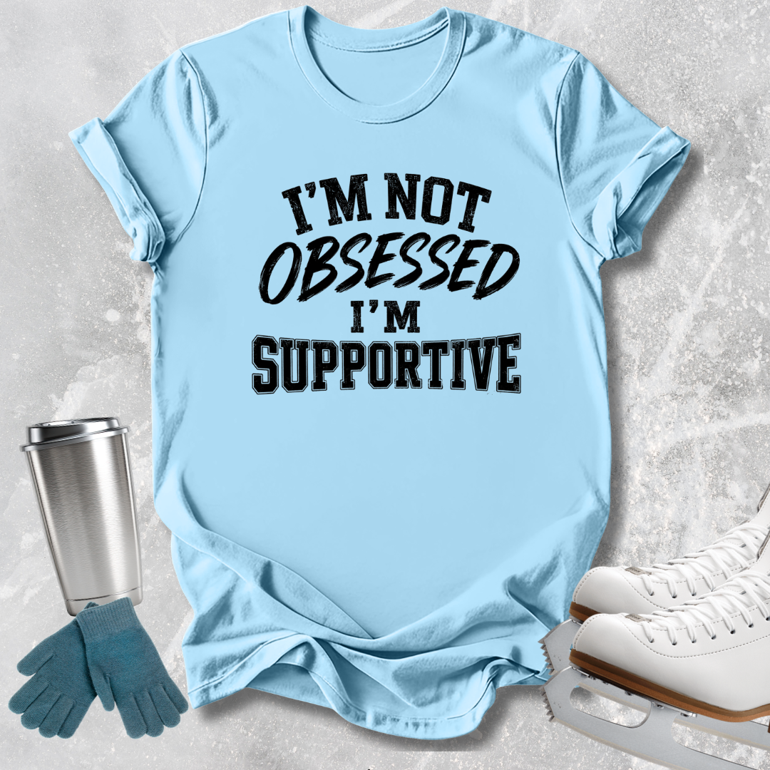 A light blue graphic t-shirt with the text 'I'M NOT OBSESSED I'M SUPPORTIVE' in black, styled in a flat-lay with ice skates, gloves, and a tumbler on a gray textured background.