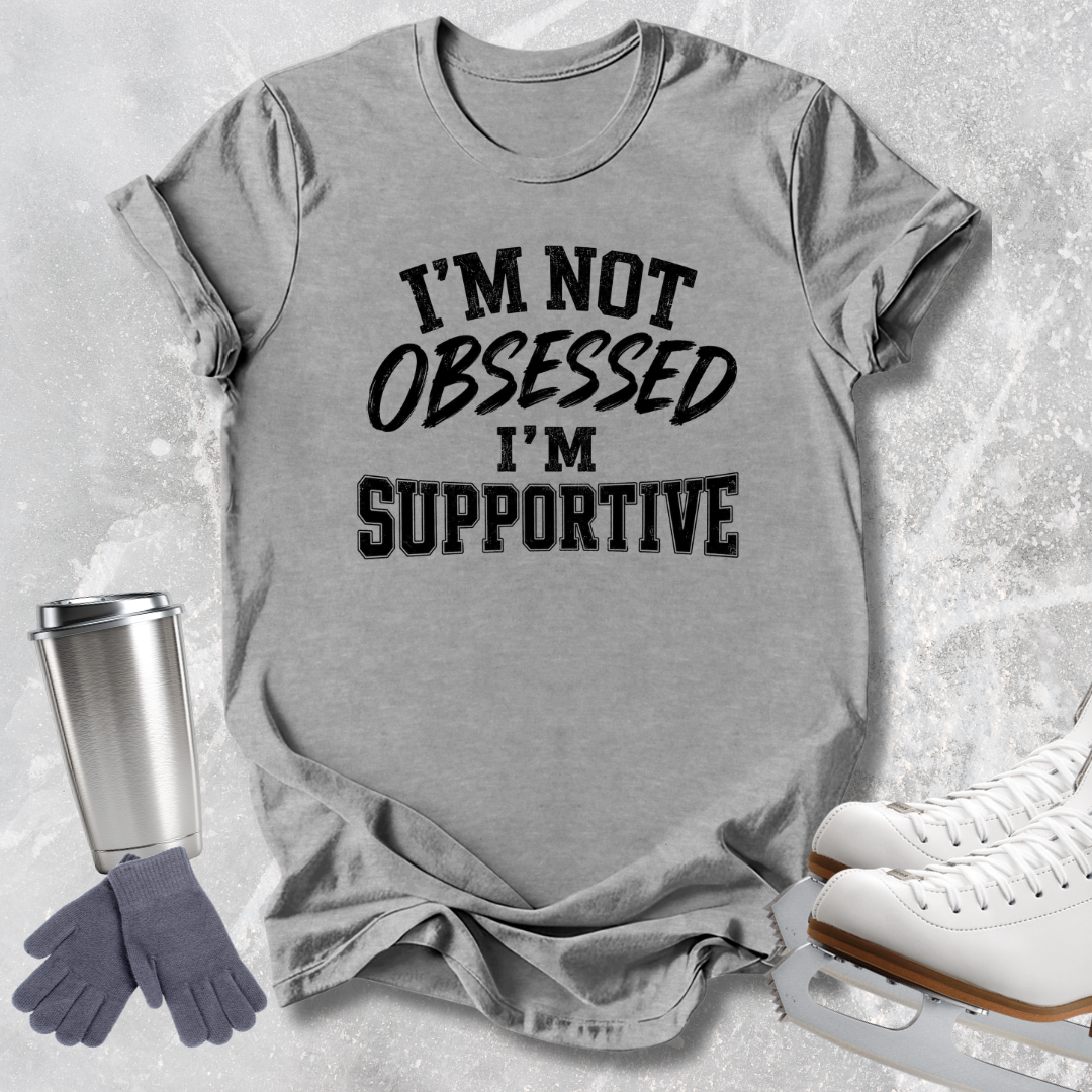 A heather gray short-sleeve t-shirt with the text 'I'M NOT OBSESSED I'M SUPPORTIVE' printed in black across the front. The shirt is styled in a flat-lay arrangement on a light gray textured background, accompanied by a silver travel tumbler, gray gloves, and white ice skates.