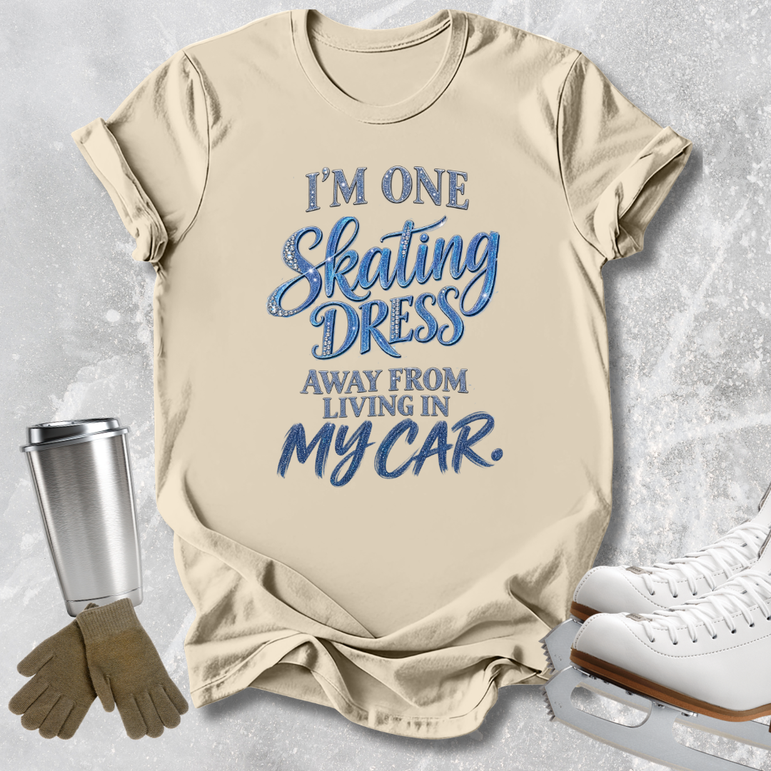 A beige graphic T-shirt laid flat on a light gray textured background, with decorative skating props including a metal cup, gloves, and ice skates. The shirt reads, 'I'M ONE Skating DRESS AWAY FROM LIVING IN MY CAR.