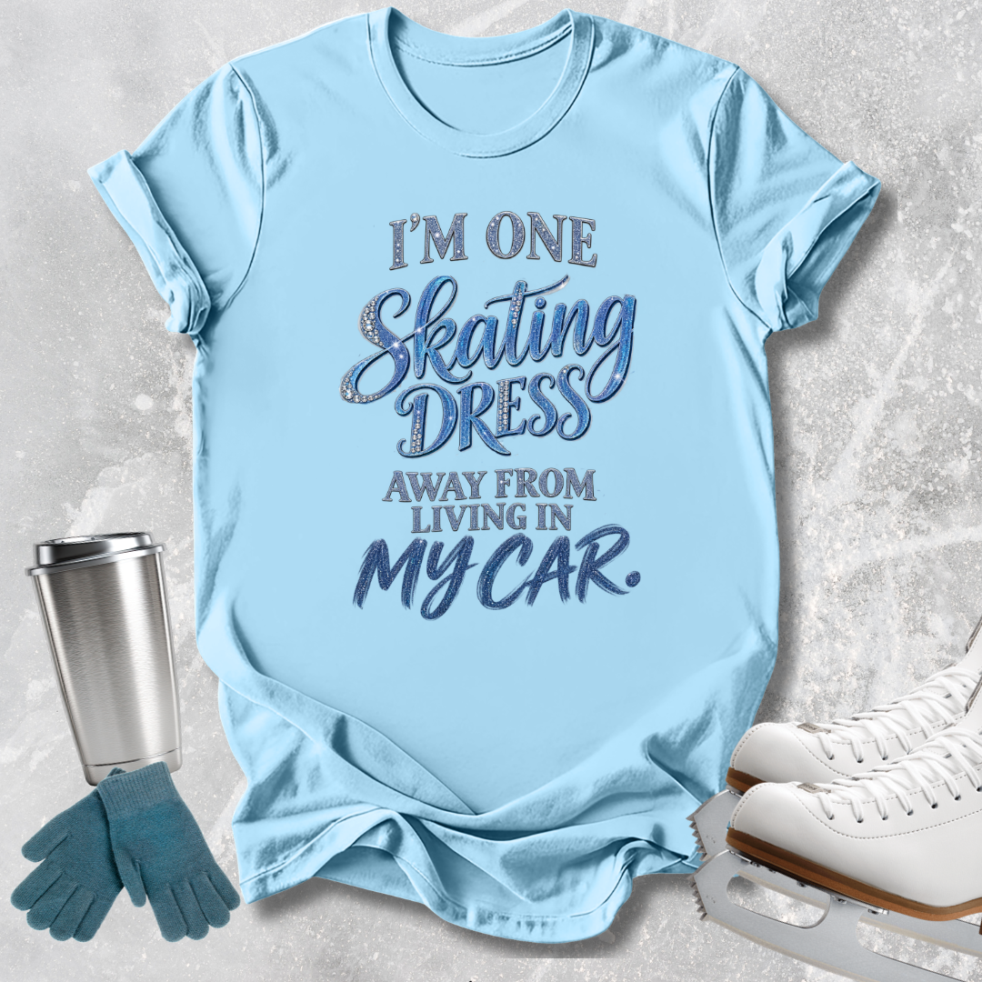 A light blue graphic t-shirt laid flat on a textured gray background, styled with ice skates, gloves, and a travel tumbler. The t-shirt features the text 'I'M ONE Skating DRESS AWAY FROM LIVING IN MY CAR.