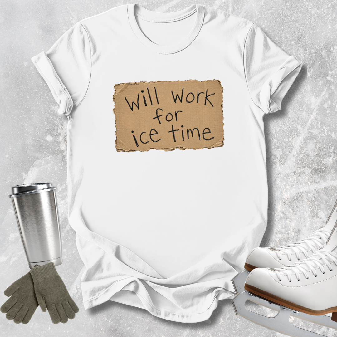 A white short-sleeve T-shirt laid flat on a light gray textured surface, featuring a tan torn-paper style graphic with the phrase 'Will Work for Ice Time' in black script. A stainless steel travel mug, gray gloves, and white ice skates are arranged around the shirt as styled props.