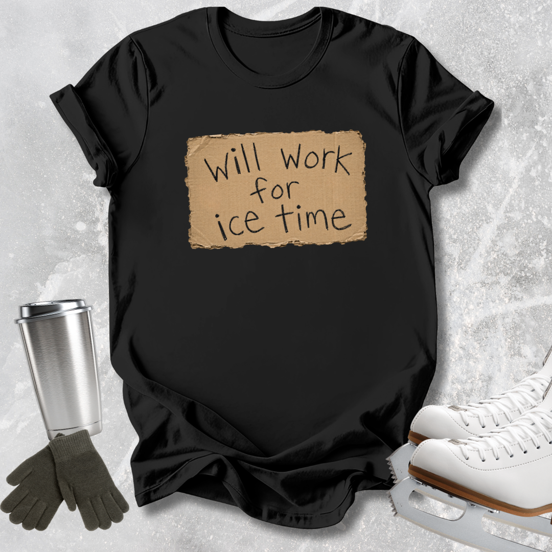 A black short-sleeve T-shirt laid flat on a light gray textured surface, featuring a tan rectangular patch with the handwritten-style text 'Will work for ice time.' Ice skates, a metal travel mug, and a pair of gloves are arranged around the shirt as styling props.