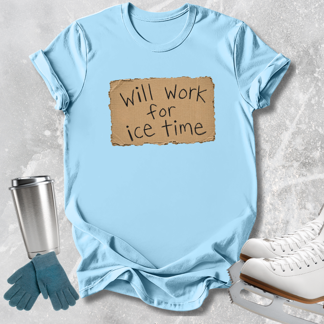 A light blue graphic t-shirt laid flat on a textured gray background, featuring a brown cardboard sign graphic that reads 'Will Work for ice time'. Ice skating accessories are styled around the shirt, including white ice skates, a silver travel mug, and blue gloves.