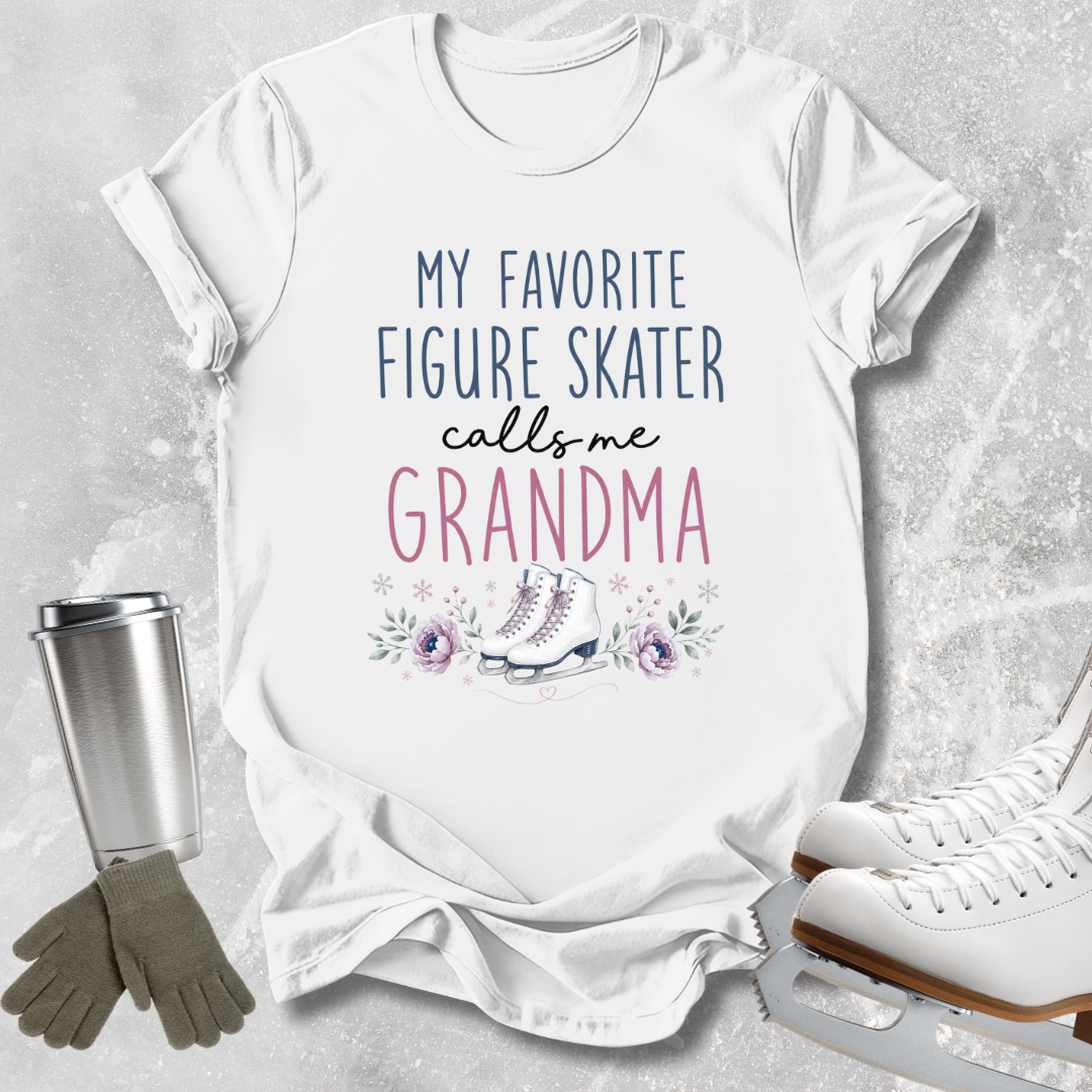 A white graphic t-shirt laid flat on a light gray textured background, featuring the text 'MY FAVORITE FIGURE SKATER calls me GRANDMA' in navy and pink lettering with a small ice skate and floral illustration. A stainless steel travel mug, gray gloves, and white ice skates are arranged around the shirt as props.