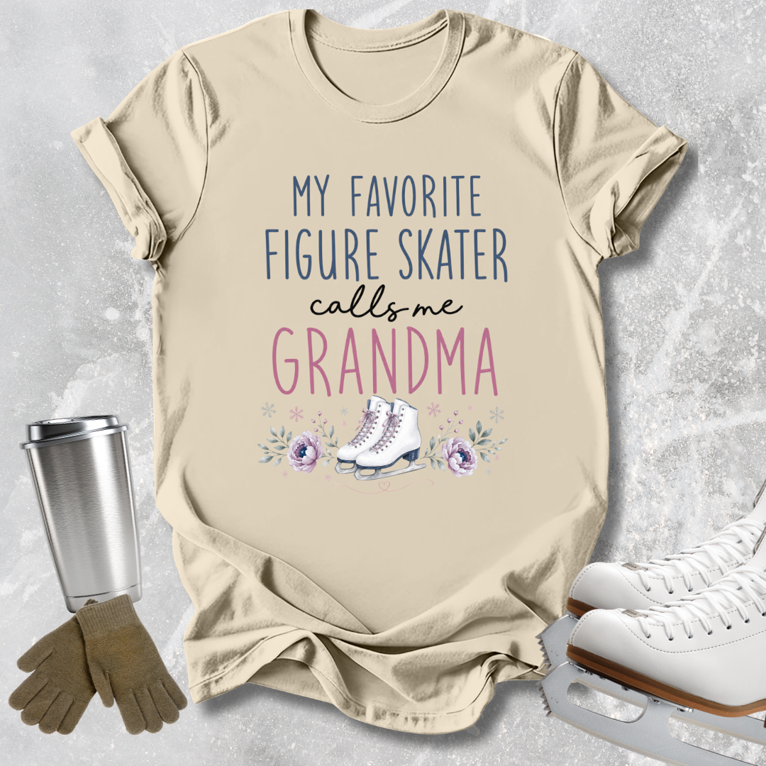 A beige graphic t-shirt featuring the text 'MY FAVORITE FIGURE SKATER calls me GRANDMA' in blue and pink lettering, decorated with a watercolor illustration of a pair of white ice skates and purple flowers. The t-shirt is styled in a flat-lay arrangement with a stainless steel travel mug, a pair of brown gloves, and white ice skates on a textured gray background.