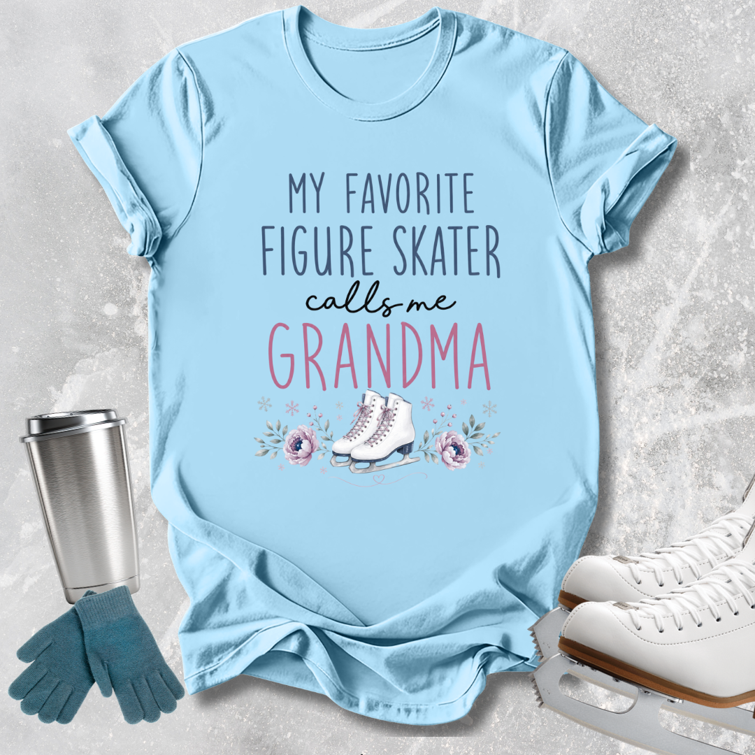 A light blue graphic t-shirt laid flat on a textured gray background, featuring the text 'MY FAVORITE FIGURE SKATER calls me GRANDMA' with a small ice skater illustration. A stainless steel tumbler, blue gloves, and white ice skates are styled around the shirt.