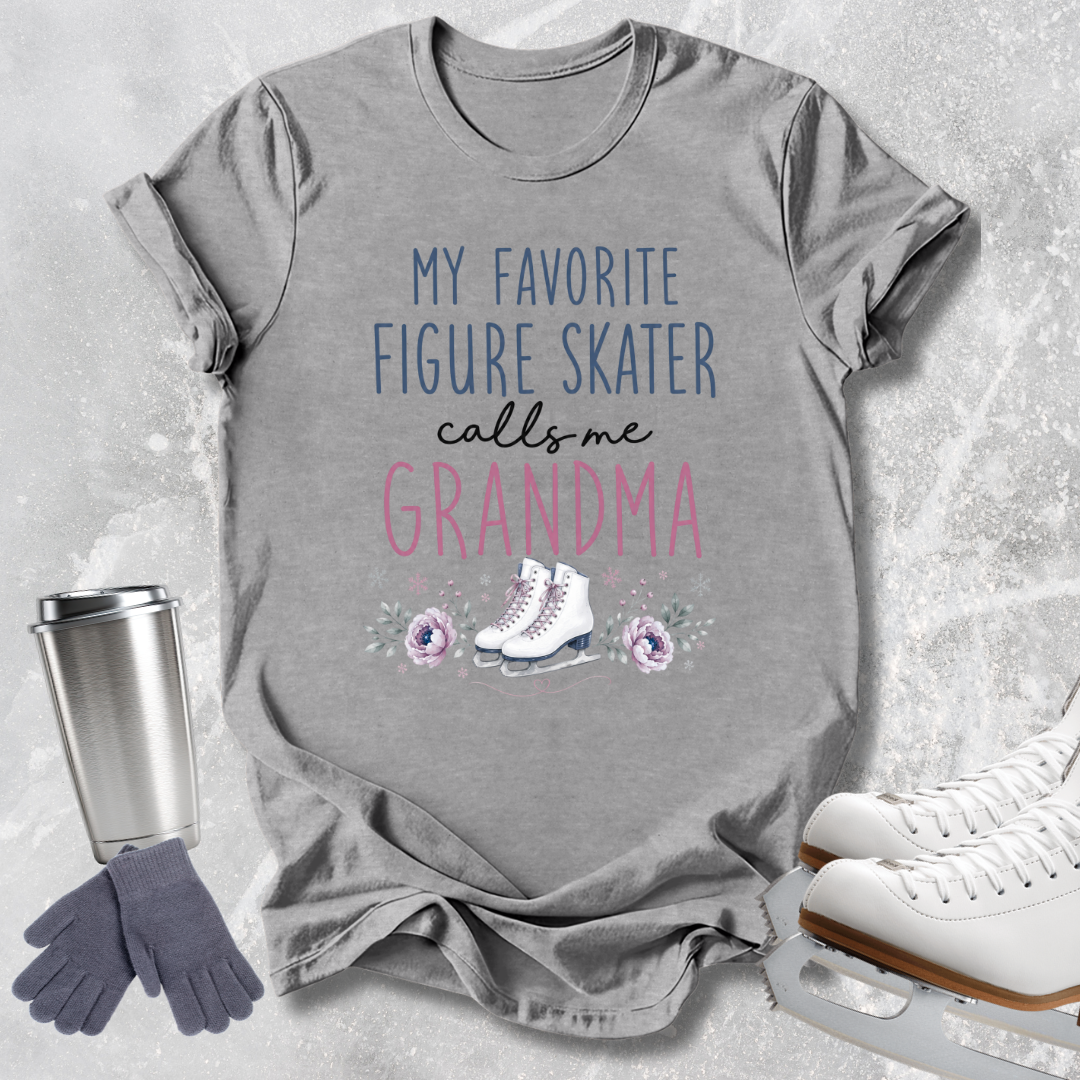 A heather gray graphic t-shirt laid flat on a light gray textured background, featuring the text 'MY FAVORITE FIGURE SKATER calls me GRANDMA' in blue and pink lettering with a small ice skater and floral illustration. A stainless steel travel mug, gray gloves, and white ice skates are arranged around the shirt as props.