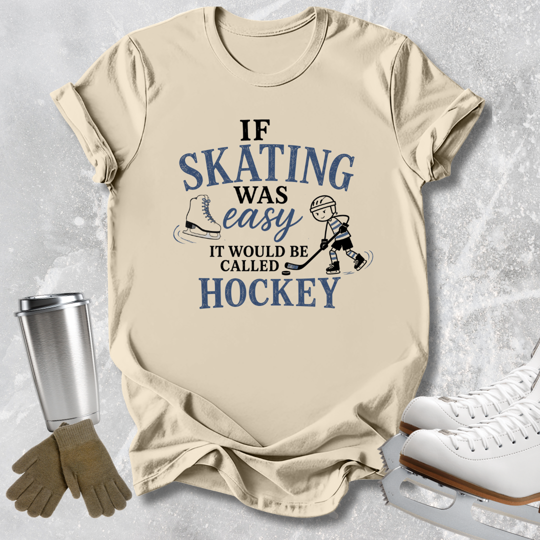 A beige graphic t-shirt featuring a blue and black hockey-themed slogan, 'IF SKATING WAS easy IT WOULD BE CALLED HOCKEY', styled in a flat-lay with ice skates, gloves, and a travel tumbler on a textured gray background.