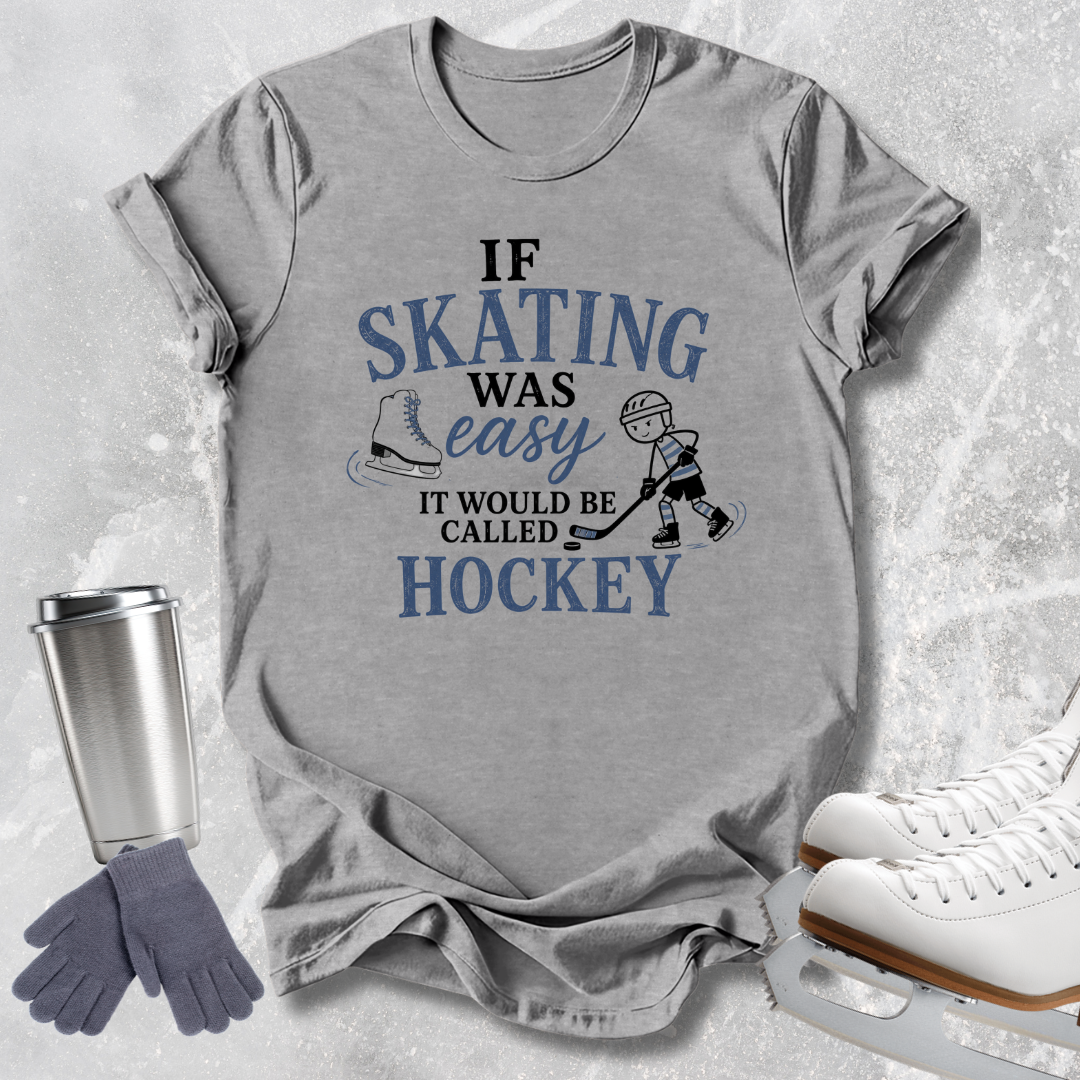 A heather gray graphic t-shirt laid flat on a light gray textured background, featuring a blue and black print that reads 'IF SKATING WAS easy IT WOULD BE CALLED HOCKEY' with skates and a hockey player illustration. A stainless steel travel mug, gray gloves, and white ice skates are arranged around the shirt as styled props.