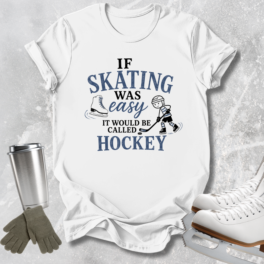 A white short-sleeve graphic T-shirt laid flat on a light gray textured surface, with skating-themed props around it: a metal cup, gloves, and ice skates. The shirt has a blue and black printed slogan reading, 'IF SKATING WAS easy IT WOULD BE CALLED HOCKEY' with skates and a player illustration.