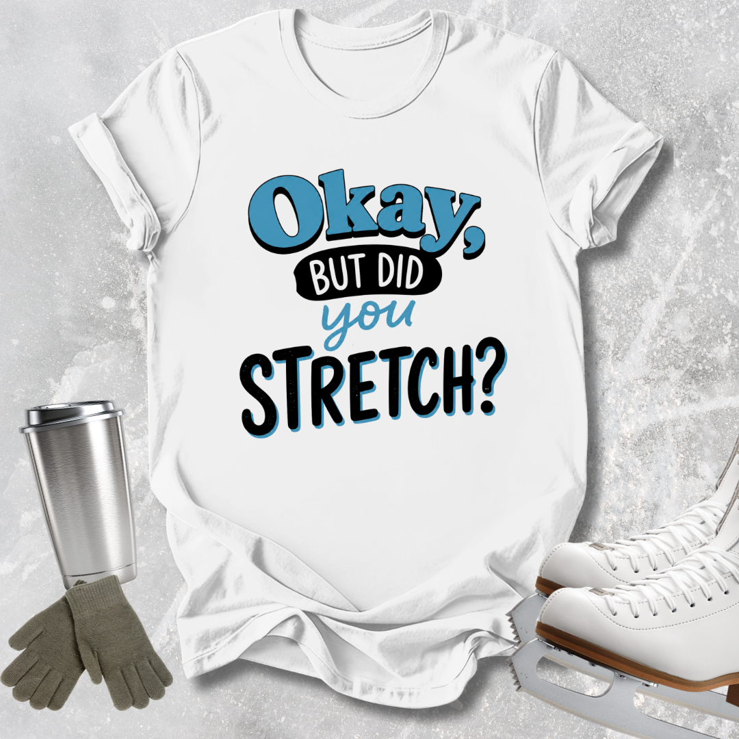 A white graphic t-shirt with the text 'Okay, BUT DID you STRETCH?' in blue and black, styled in a flat lay with ice skates, gloves, and a metal cup on a textured gray background.