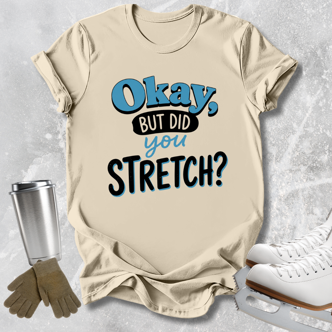 A beige graphic t-shirt with the text 'Okay, BUT DID you STRETCH?' in blue and black, styled in a flat lay with ice skates, gloves, and a travel tumbler on a gray textured background.