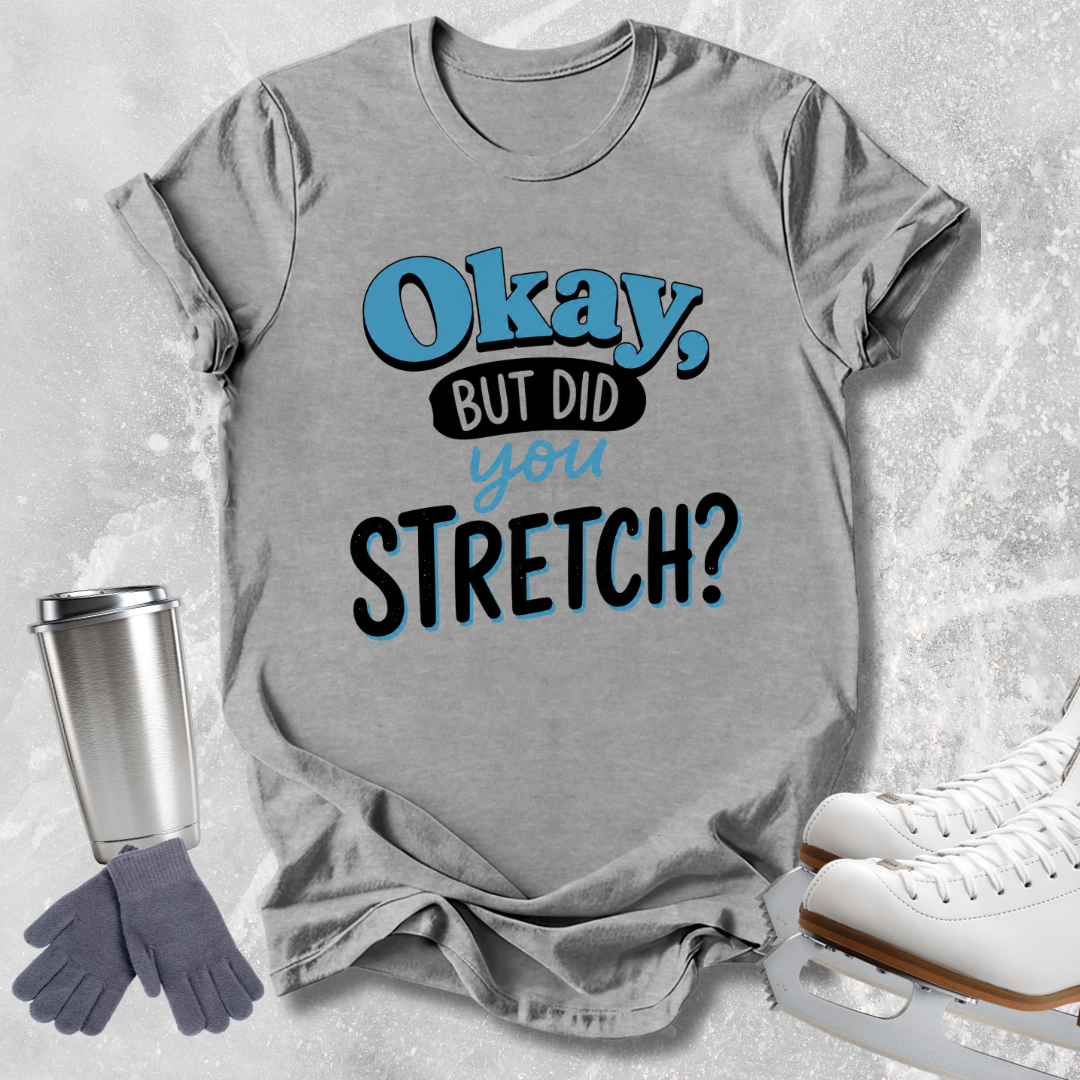 A heather gray graphic T-shirt laid flat on a light textured background, with the phrase 'Okay, BUT DID you STRETCH?' printed in blue and black. Ice skates, gloves, and a travel tumbler are arranged around the shirt as styling props.