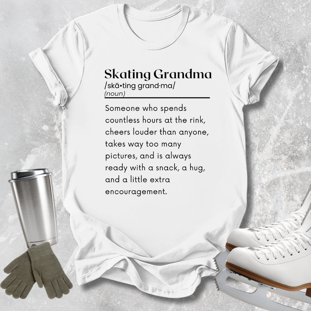 A white graphic T-shirt laid flat on a light gray textured surface, with a printed definition of the phrase 'Skating Grandma' in black text. Surrounding props include a metal travel mug, gray gloves, and white ice skates.