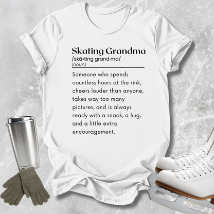 A white graphic T-shirt laid flat on a light gray textured surface, with a printed definition of the phrase 'Skating Grandma' in black text. Surrounding props include a metal travel mug, gray gloves, and white ice skates.
