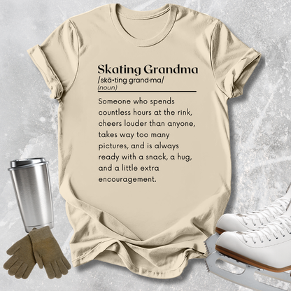 A beige graphic T-shirt laid flat on a light gray textured surface, with skating-themed props including white ice skates, gloves, and a metal cup. The shirt reads 'Skating Grandma' with a dictionary-style definition printed in black text.