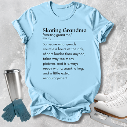 A light blue graphic T-shirt laid flat on a light gray textured surface, with a printed dictionary-style definition reading 'Skating Grandma.' Surrounding props include a metal travel tumbler, blue gloves, and white ice skates.