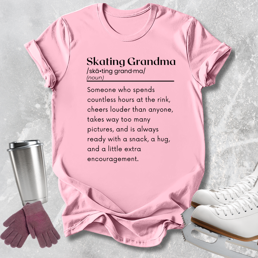 A light pink graphic T-shirt laid flat on a textured light gray background, with a stainless steel cup, gloves, and white ice skates around it. The shirt has black printed text defining 'Skating Grandma.