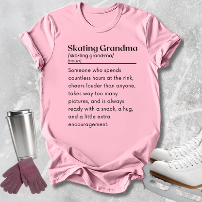 A light pink graphic T-shirt laid flat on a textured light gray background, with a stainless steel cup, gloves, and white ice skates around it. The shirt has black printed text defining 'Skating Grandma.