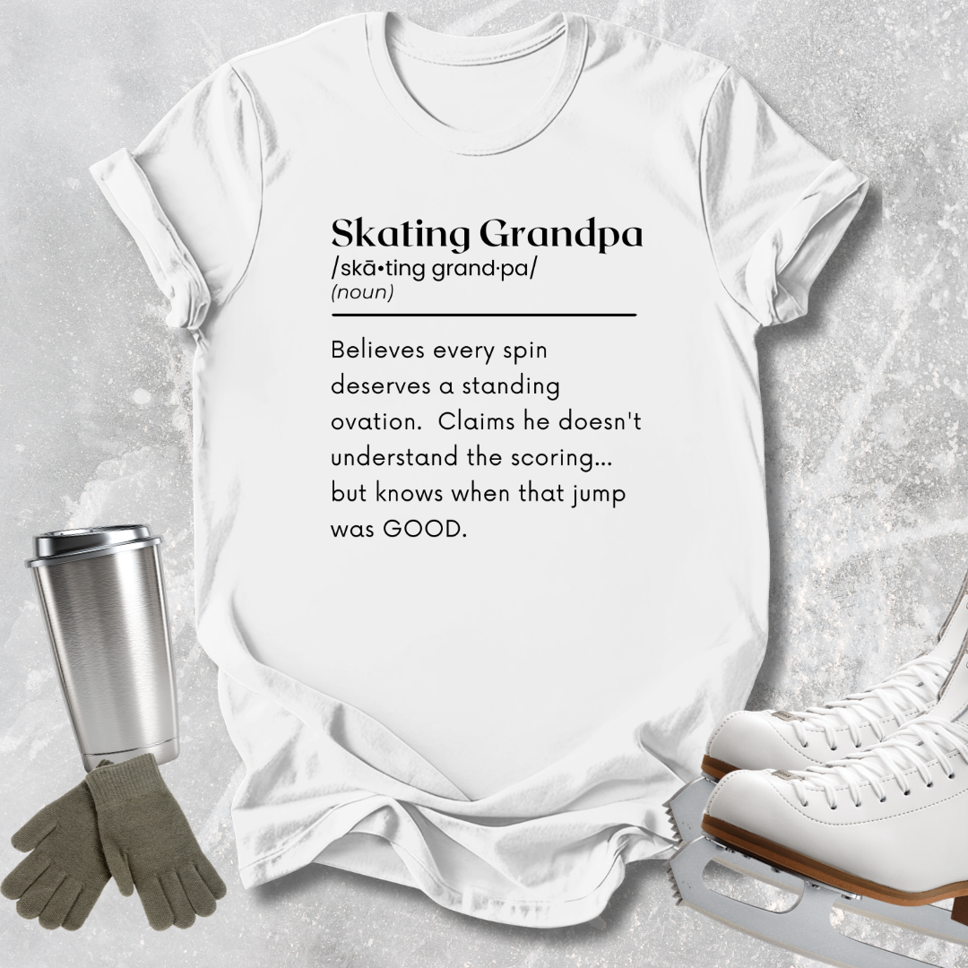 A white graphic T-shirt laid flat on a light gray textured surface, with printed black text defining 'Skating Grandpa.' Ice skates, gloves, and a metal cup are arranged around the shirt as styling props.