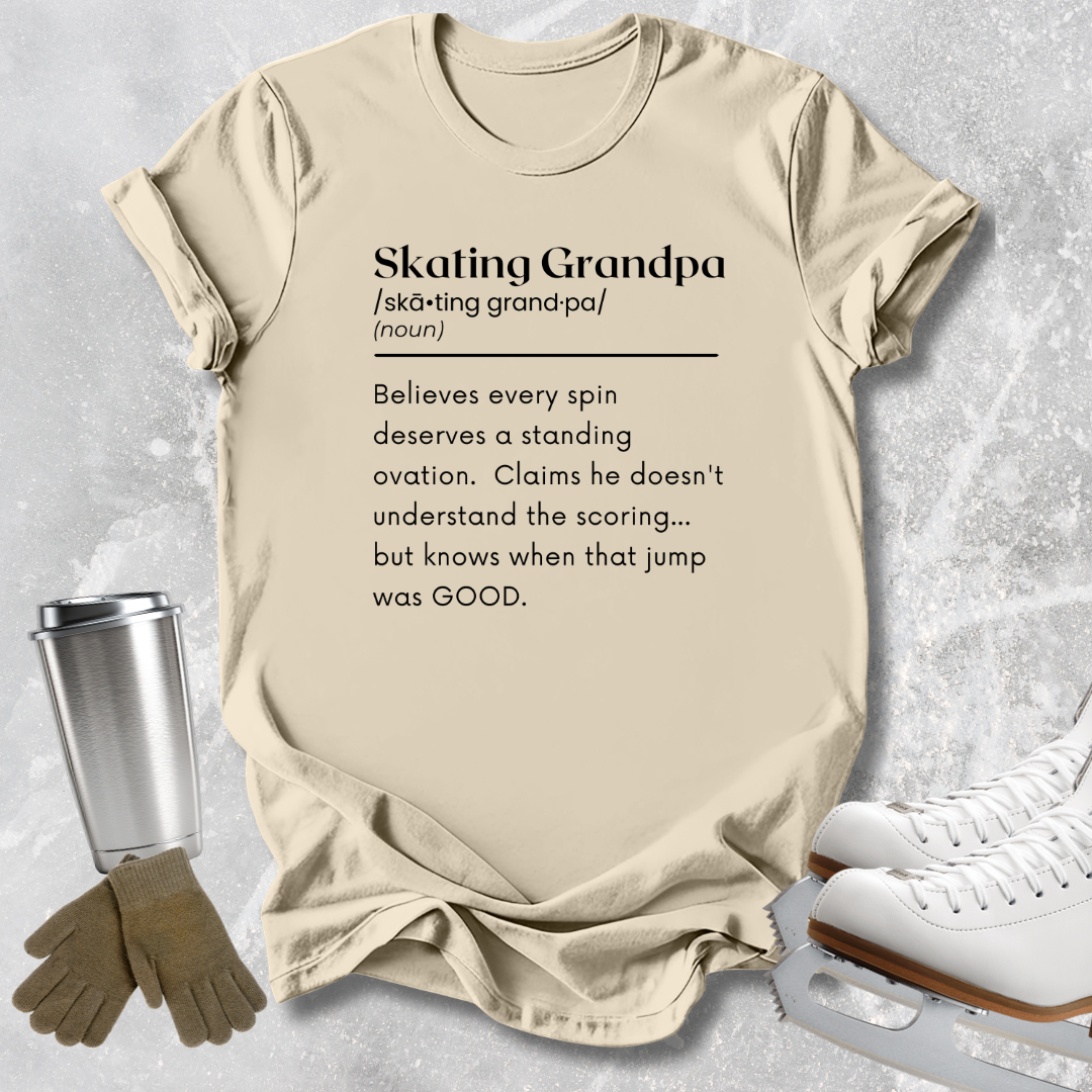 A beige graphic t-shirt featuring a humorous dictionary definition of 'Skating Grandpa' printed in black text. The t-shirt is styled in a flat-lay arrangement on a textured gray background, accompanied by a silver travel mug, a pair of brown gloves, and white ice skates.