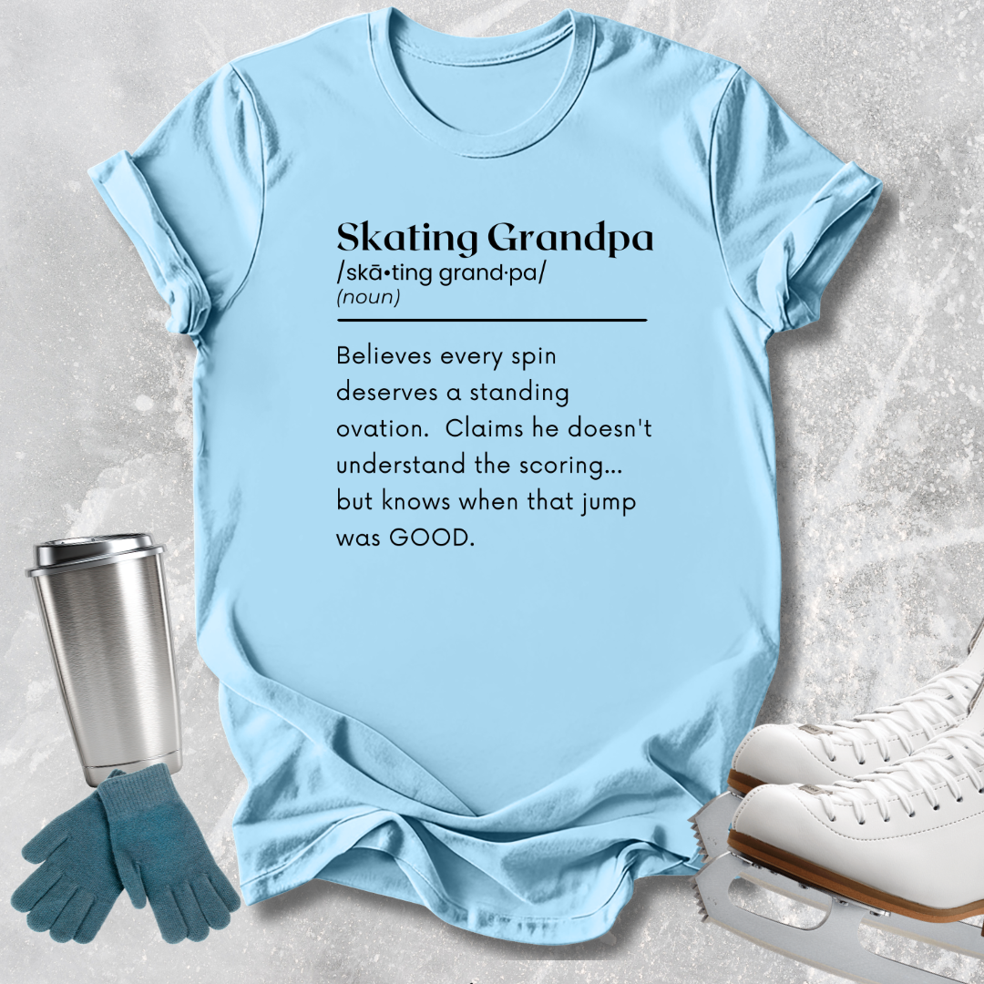 A light blue graphic T-shirt laid flat on a textured light gray surface, with printed black text defining 'Skating Grandpa.' White ice skates, teal gloves, and a stainless steel tumbler are arranged around it as styling props.
