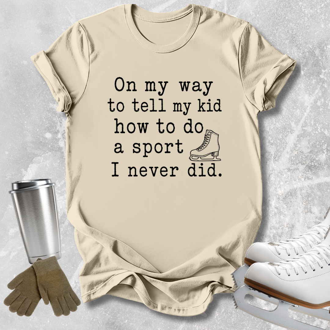 A beige graphic t-shirt laid flat on a light gray textured background, with black text reading 'On my way to tell my kid how to do a sport I never did.' and a small ice skate illustration.